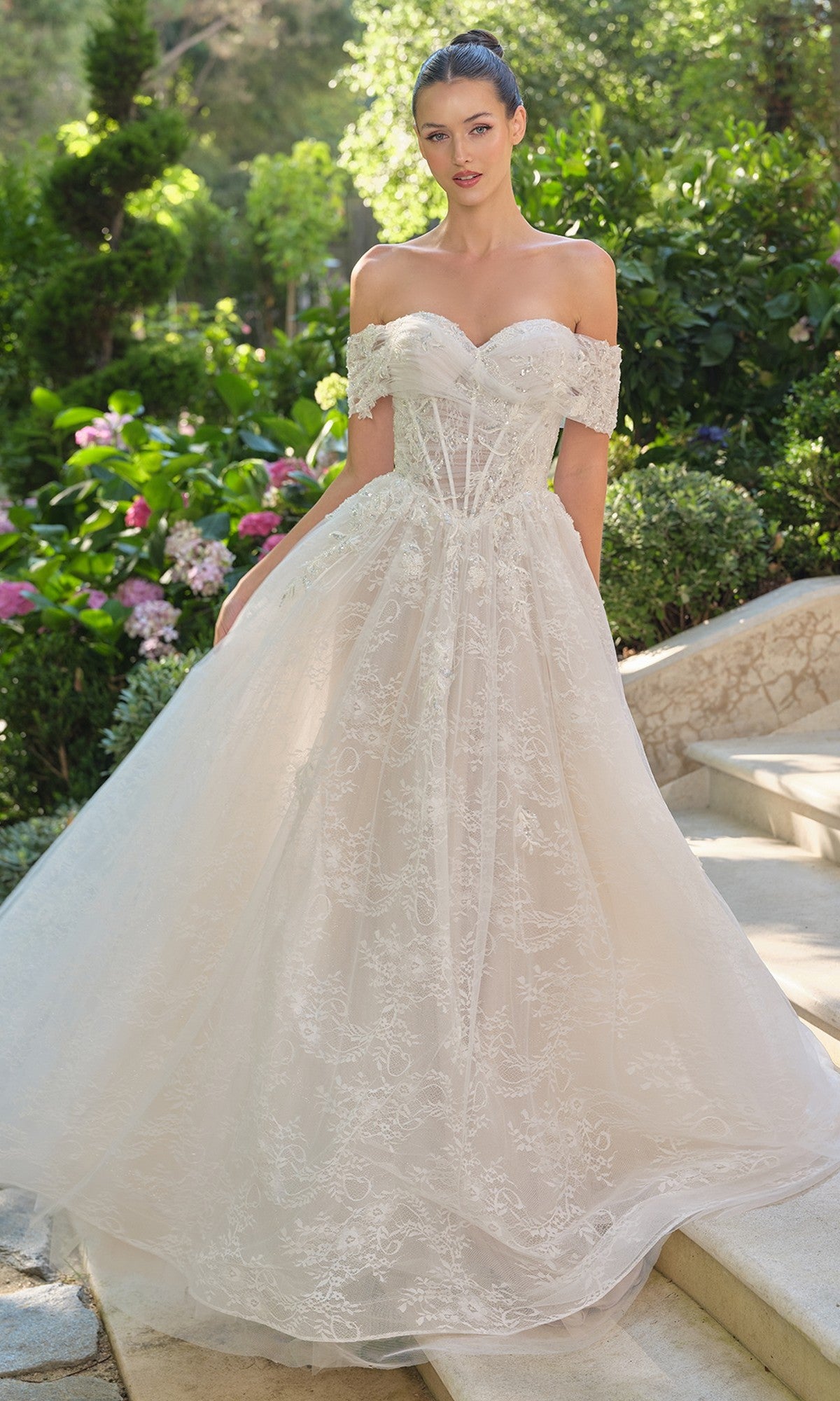 A woman stands outdoors in the Andrea and Leo WL090 long wedding dress, featuring an off-the-shoulder lace design with a corset bodice and full skirt, surrounded by lush greenery and blooming flowers.
